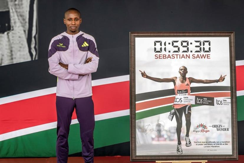 Kenyan marathon runner Sabastian Sawe poses on stage next to a picture of him while crossing the finish line to win the London Marathon after breaking a historic world record, during a congratulatory ceremony hosted by Kenya's President William Ruto (unseen) at State House in Nairobi, on April 30, 2026. Sabastian Sawe became the first man to run under two hours for a marathon in race conditions thanks to extraordinary talent, a punishing training regime, and the lightest shoes in history. In London on April 26, 2026, the 31-year-old Kenyan achieved one of sport's milestones, ranking alongside Roger Bannister's first sub-four minute mile in 1954. Luis TATO / AFP